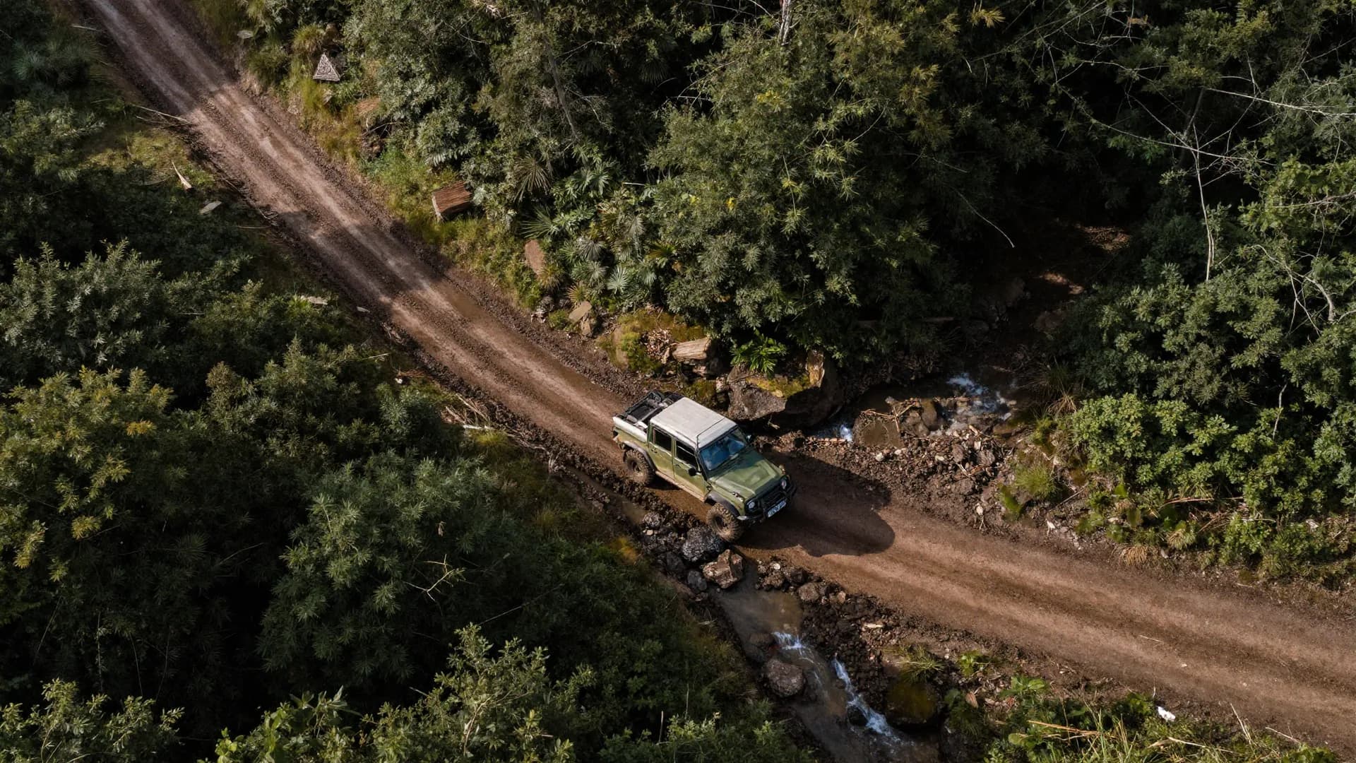 Top-down aerial drone shot of a winding muddy trail through forested terrain with a single off-road truck at a creek crossing